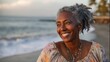 © mila103 - A joyful woman with grey curls stands on the sandy beach, smiling softly while gazing at the waves. The sun sets behind her, casting a warm glow on the tranquil scene.