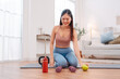 © Kiattisak - Woman taking a break after home workout, sitting on yoga mat with dumbbells, kettlebell, green apple, and water bottle. Healthy lifestyle, post-exercise recovery, and fitness nutrition at home.