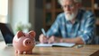 © Maryna - Elderly man with white beard uses laptop for accounting and finances. Pink piggy bank sits on desk next to notebook for savings and budget planning. He is writing in planner.