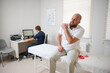 © Василь Івасюк - Male patient being examined in clinic for shoulder pain. Male suffering from shoulder pain. Patient sitting on examination couch holding his arm while doctor works at computer in background.