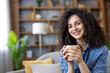 © Liubomir - Smiling woman with curly brown hair relaxing on a cozy sofa in her modern living room, holding a warm mug and enjoying a peaceful morning break, content and comfortable