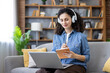 © Liubomir - Young woman with curly hair wearing headphones and taking notes in a notebook, while sitting on a sofa and using a laptop for online learning or remote work at home