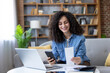© Liubomir - Woman sitting on a sofa, happily reviewing paperwork, holding a smartphone, and using a laptop to manage her budgeting, banking, and online bill payments at home