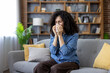 © Liubomir - Young woman with curly hair sitting on a sofa in a living room, blowing her nose with a tissue feeling sick, showing symptoms of a cold, flu, virus, or seasonal allergies