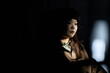 © TrueFrame Collective - Asian young adult woman sitting in partial shadow, looking down thoughtfully, dark hair visible, appearing deep in contemplation, soft light highlighting face