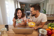 © Stockphotodirectors - A couple shares a joyful moment over breakfast in their warm kitchen. They are eating from colorful bowls and looking at a laptop, smiling and engaging with each other's company.