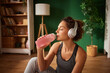 © Stockphotodirectors - A woman sits on the floor, sipping a pink drink from a bottle while wearing headphones. The room features natural light and a green wall, creating a peaceful atmosphere.