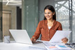 © Liubomir - Indian businesswoman wearing glasses sitting at an office desk, smiling while working on a laptop and holding documents, focusing on business, finance, and career growth