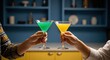 © Mr. Stocker - Close-up of two people toasting with colorful green and yellow martini cocktails, celebrating together in a stylish kitchen with blue and yellow decor