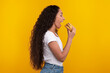 © Prostock-studio - A smiling woman holds a tasty burger in her hand, preparing to take a big bite. She has long curly hair and wears a casual outfit against a vibrant yellow background.