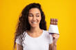 © Prostock-studio - A cheerful woman with curly hair stands in a vibrant yellow studio, holding a chocolate bar. She smiles while biting her lip, showcasing her love for sweets and the joy of indulging.