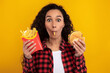 © Prostock-studio - A joyful lady is having a fun time eating a burger and french fries, holding a piece of potato in her mouth. The vibrant yellow background adds to the playful mood of this fast food moment.