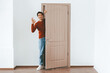 © Prostock-studio - A young man stands next to an open door, smiling and gesturing okay with his hand. The setting is bright and minimalistic, featuring light colored walls and wooden flooring.