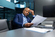 © Anastasiya - Serious mature African American businessman analyzing documents in modern office. Focused corporate leader reviewing paperwork with a thoughtful, concentrated expression