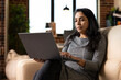 © DC Studio - Modern Indian woman seated on couch, focused on editing blog post and researching business trends. Female entrepreneur working remotely with laptop, reviews marketing plans in cozy home environment.