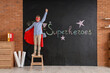 © Pixel-Shot - Teenage boy dressed as superhero on stepladder near blackboard in classroom