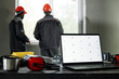 © Seventyfour - Two middle aged men wearing safety helmets standing near window discussing construction project, foreground showing open laptop with blank screen and protective headphones