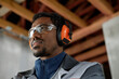 © Seventyfour - Portrait of young adult Black man wearing protective earmuffs and safety glasses working in industrial setting, looking focused and attentive, visible wooden beams in background