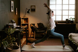 biracial young adult woman practicing yoga in living room wearing electronic ankle monitor, stretching arms upward while standing on exercise mat near window during home confinement