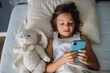 © Anna Lurye - Young girl lying in bed with her plush toy, using a smartphone, illustrating modern childhood, screen time, and children's engagement with technology