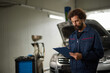 © Stockphotodirectors - A car mechanic is focused on evaluating the condition of a vehicle in a repair shop. He is checking a clipboard filled with notes, surrounded by tools and equipment.