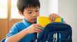 © Nii_Anna - Asian boy packing school bag with books in classroom setting