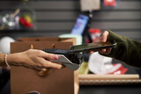 Caucasian woman hand holding payment terminal while young adult hand holding smartphone making contactless payment in retail store with shopping bags visible in background