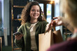 © DragonImages - Young adult Caucasian woman smiling while receiving shopping bag from another , holding smartphone in hand, standing indoors in modern retail environment