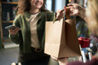 © DragonImages - Young adult Caucasian woman smiling while holding smartphone and receiving paper shopping bag from middle aged Caucasian woman during transaction at flower shop counter