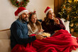 © Home-stock - Happy family with daughter watching Christmas movie, eating popcorn while lying under blanket on sofa in living room with tree and glowing garlands