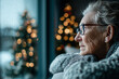 © Olha Havelia - elderly woman sitting alone in her living room, looking out the window at the christmas tree and lights outside, feeling lonely during the holiday season. copy space. high-quality,