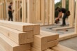 © hobonski - Smooth timber beams lie neatly stacked in the foreground while workers assemble a wooden frame behind them, showing progress, structure, and early construction stages.