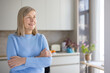 © Liubomir - Senior woman looking away smiling, standing with arms crossed in her bright kitchen, experiencing happiness and positive emotions in her comfortable home