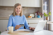 © Liubomir - Smiling senior woman in a cozy kitchen using a laptop and holding a credit card for online shopping and banking, relaxed at the counter with a mug and natural light