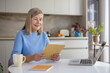 © Liubomir - Senior woman smiling while unpacking and reading a letter or important document at a laptop-equipped table, managing paperwork at her home office kitchen