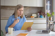 © Liubomir - Senior woman feeling chest pain and stress while reading an upsetting document from an envelope, sitting at a table in her kitchen with a laptop and coffee