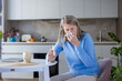 © Liubomir - Senior woman sitting at a table in the kitchen, blowing her nose with a tissue and holding a nasal spray, experiencing seasonal allergies, flu, or illness
