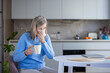 © Liubomir - Senior woman suffering from a cold, flu, or allergy symptoms, holding a mug and blowing her nose with a tissue while feeling unwell at home in the kitchen