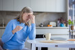 © Liubomir - Mature woman sitting at a kitchen table, rubbing her temples and holding eyeglasses, expressing discomfort and pain from a severe headache, reflecting stress and fatigue