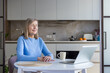 © Liubomir - Senior woman in blue sweater meditating with eyes closed at a bright kitchen table, laptop and coffee nearby, practicing mindfulness, calm and balance during remote work
