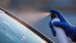 ©  VladaToday - Hand in blue glove spraying deicer onto car windshield to remove ice with visible droplets and partial view of vehicle