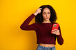 © deagreez - Young mixed race woman against yellow background wearing a maroon sweater and checks her smartphone with puzzled expression while shopping