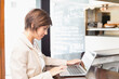 © wavebreak3 - Woman in 20s-30s sitting at cafe counter, typing on laptop by chalkboard menu, wearing beige blazer