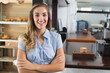 © wavebreak3 - Female barista standing at counter in light blue blouse, showing wooden shelves and coffee grinder