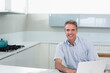 © wavebreak3 - Middle-aged man in blue shirt sitting at kitchen counter using laptop beside teal pot, copy space