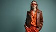 © Maryna - Stylish young woman poses in studio wearing bright orange pants with a leather jacket. Beautiful model with sunglasses. Fashionable brunette lady presents vogue outfit on gray backdrop. Modern look.