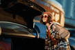 © SHOTPRIME STUDIO - Woman by car trunk with bag and sunglasses wearing beanie and plaid coat, golden hour candid lifestyle portrait capturing authenticity, mindful living and emotional storytelling glow.