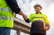 © tong2530 - Two construction engineers in safety gear shake hands, finalizing a deal or showing successful teamwork at a construction site.