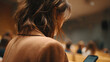 © jayantha - A student or attendee in a brown blazer checks their smartphone, captured from behind in a blurry lecture hall setting.