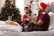 © New Africa - Happy couple in pajamas with cups of hot drink spending time together on bed in room decorated for Christmas